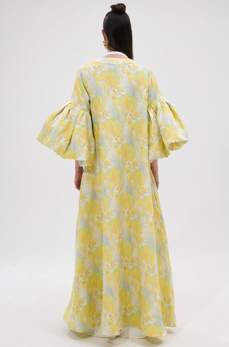 A person stands facing away, wearing the VENICE BROCADE PUFF SLEEVE DUSTER—a long, flowy dress with puff sleeves and a yellow and light blue floral brocade pattern—against a plain white background.