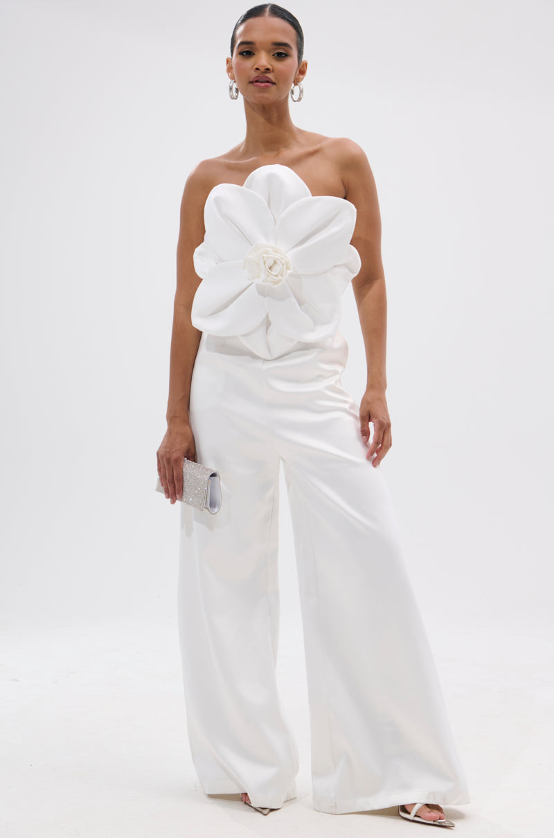 A woman stands against a white background wearing the SUMMER BLOOMS WIDE LEG JUMPSUIT IN WHITE, featuring a strapless design, wide-leg pants, and dramatic 3D floral appliqué on the bodice. She carries a silver clutch and wears hoop earrings. 0