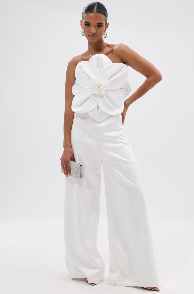A woman stands confidently in the SUMMER BLOOMS WIDE LEG JUMPSUIT IN WHITE, featuring a strapless design, 3D floral applique bodice, wide-leg pants, and holds a silver clutch against a plain white background. 1
