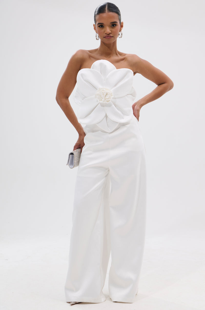 A woman stands confidently in the SUMMER BLOOMS WIDE LEG JUMPSUIT IN WHITE, featuring a strapless design, dramatic floral applique at the bodice, and wide legs. She accessorizes with a clutch and hoop earrings against a plain white background. 2