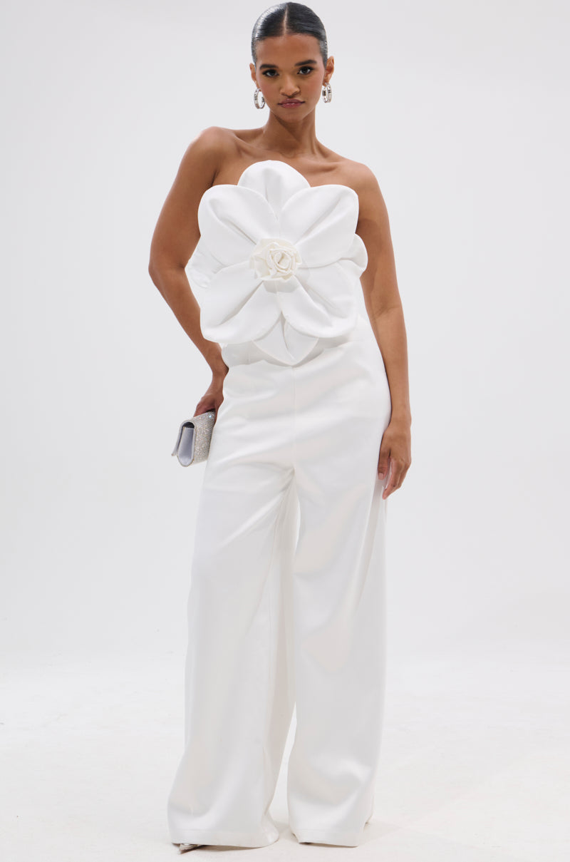 Against a plain white background, a woman models the SUMMER BLOOMS WIDE LEG JUMPSUIT IN WHITE, featuring a strapless design with a bold floral applique on the bodice. She completes the look with hoop earrings, slicked-back hair, and a small clutch. 3