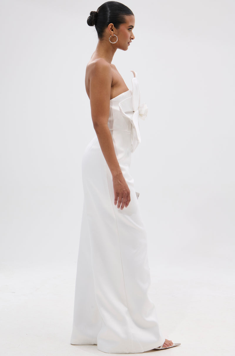 A woman in profile poses against a white background wearing the SUMMER BLOOMS WIDE LEG JUMPSUIT IN WHITE, featuring a strapless design, wide legs, and a large bow on the bodice. She styles her hair in a sleek bun with hoop earrings. 4