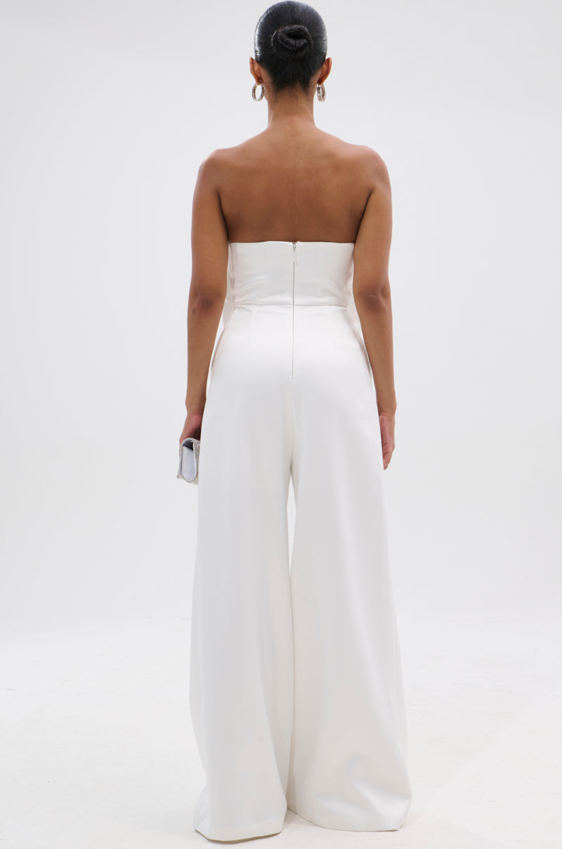 A woman with her hair in a bun is seen from behind wearing the SUMMER BLOOMS WIDE LEG JUMPSUIT IN WHITE and holding a small clutch, with a plain white background highlighting the subtle elegance of her look. 5
