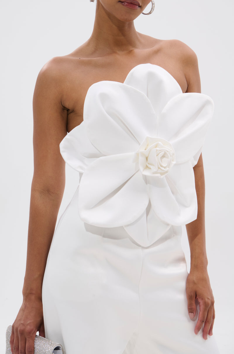 A woman models the SUMMER BLOOMS WIDE LEG JUMPSUIT IN WHITE, featuring a strapless design and 3D floral applique on the bodice, while holding a glittery clutch against a plain background. 6