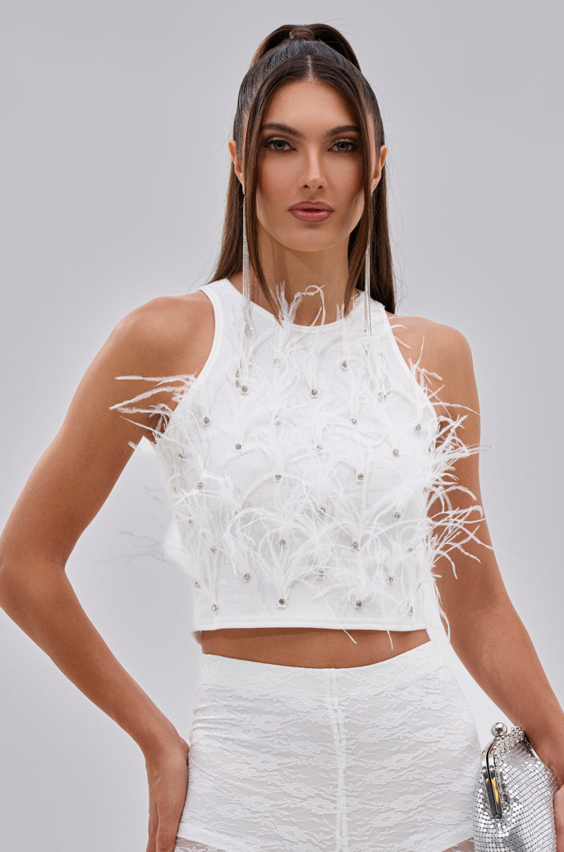 A woman with long dark hair in a high ponytail wears the BIRDS OF A FEATHER SLEEVELESS TOP with pearl accents, paired with high-waisted white lace bottoms, standing against a plain light gray background. 0