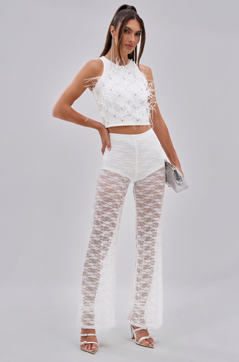A woman stands against a plain background wearing the BIRDS OF A FEATHER SLEEVELESS TOP, high-waisted sheer lace pants, white heels, and holding a silver clutch. Her straight hair features two front strands pulled forward. 7