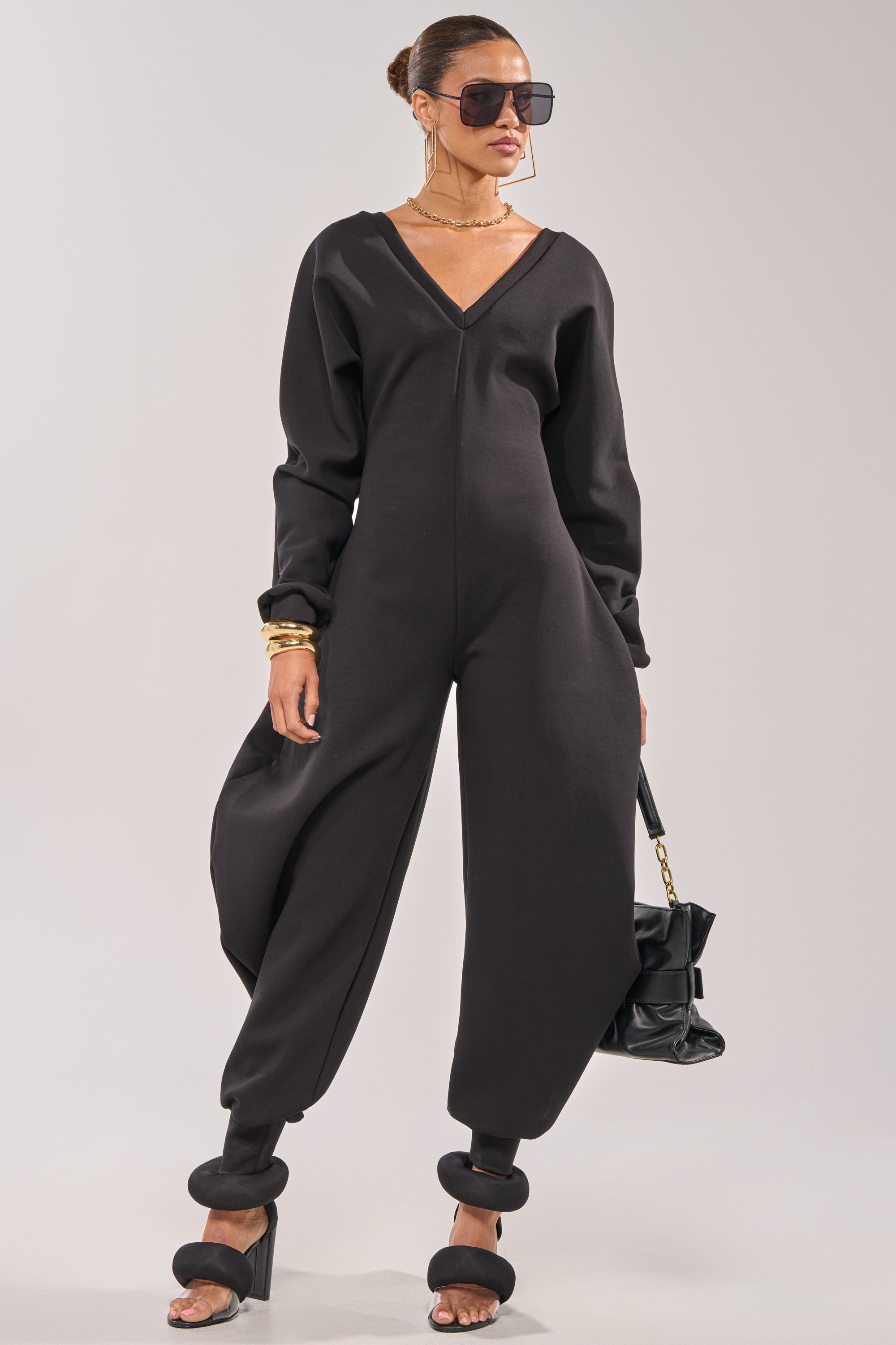A woman stands confidently holding a black handbag, wearing the COMFY COZY SWEATSHIRT JUMPSUIT IN BLACK with wide legs, oversized sunglasses, hoop earrings, bangles, and heeled sandals. 0