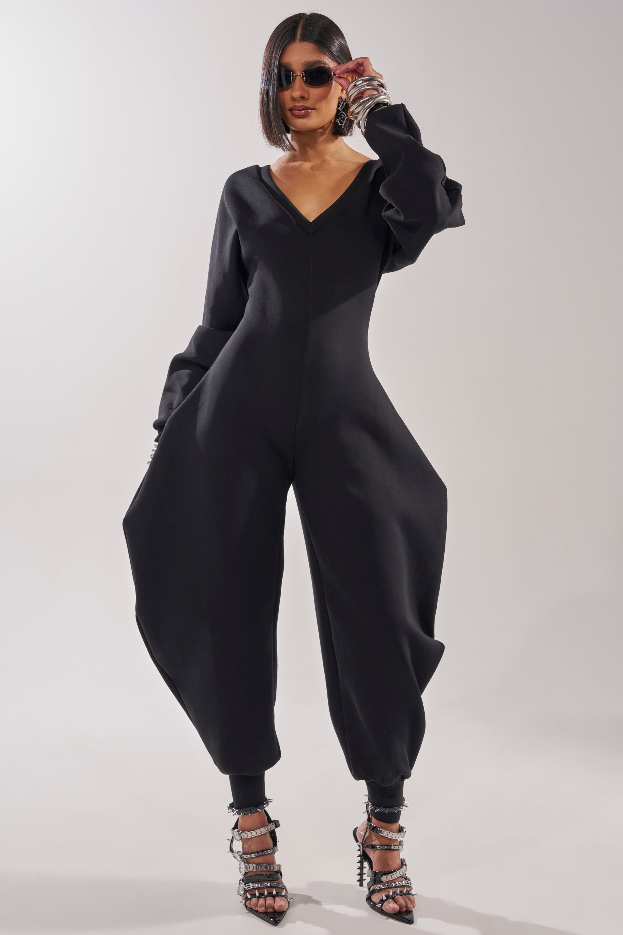 Wearing the COMFY COZY SWEATSHIRT JUMPSUIT IN BLACK, paired with dark sunglasses and strappy heeled sandals, a confident person poses against a plain light background.