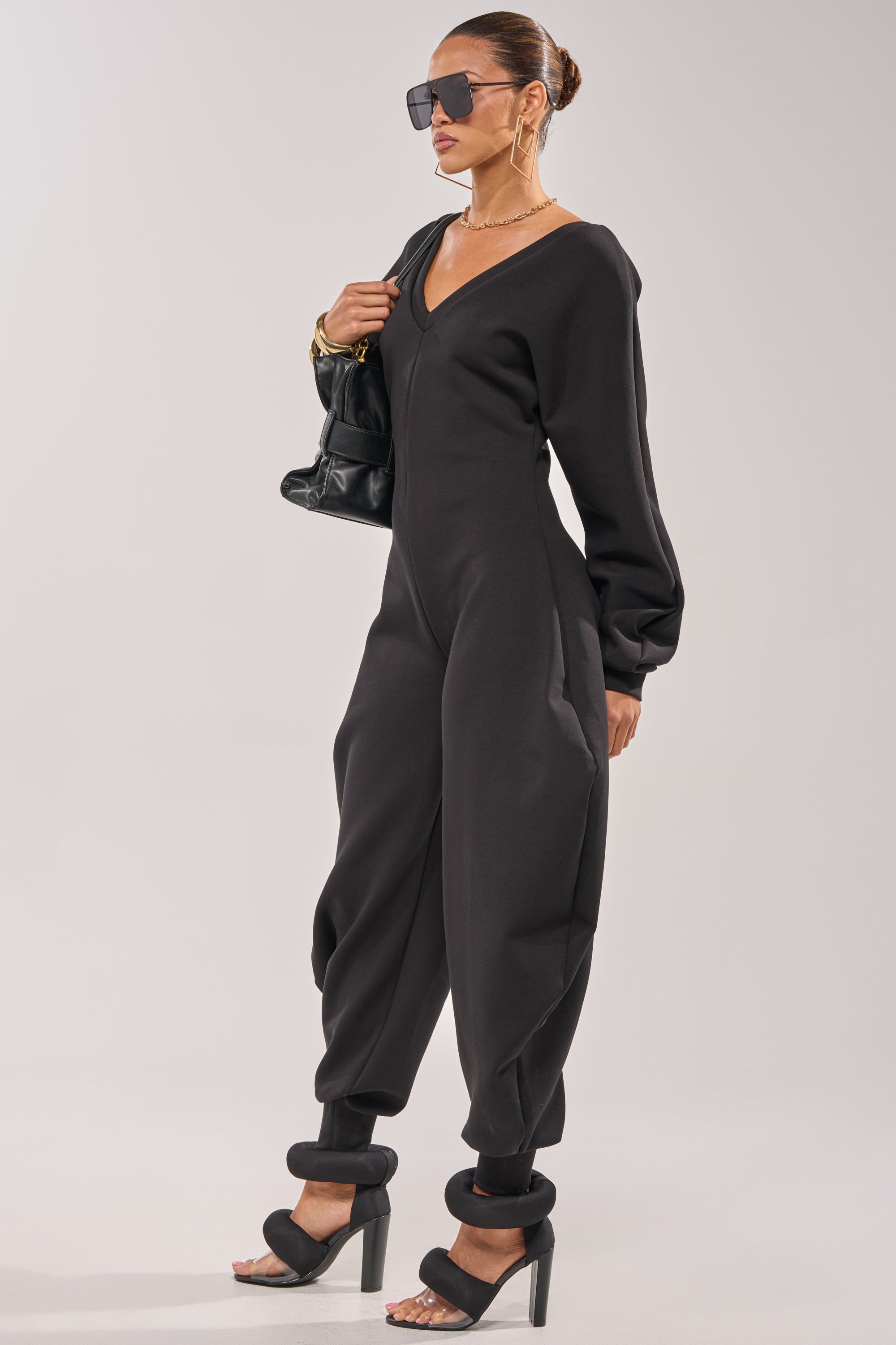 A woman stands confidently against a plain light background wearing the COMFY COZY SWEATSHIRT JUMPSUIT IN BLACK, paired with black high heel sandals, large sunglasses, gold jewelry, and a black handbag. 1