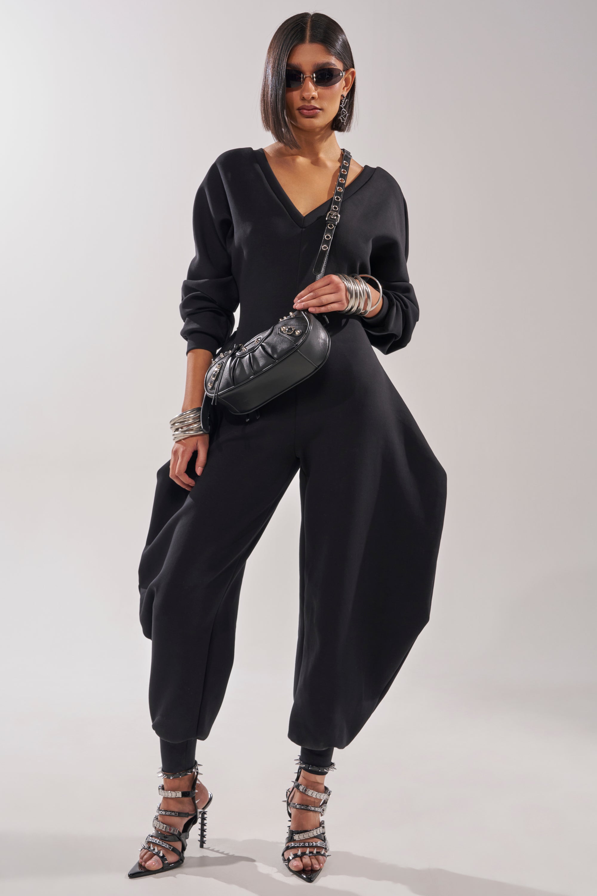 A woman with a sleek bob and sunglasses wears the COMFY COZY SWEATSHIRT JUMPSUIT IN BLACK, studded heels, chunky bracelets, and a black crossbody bag, standing confidently against a plain background. 1
