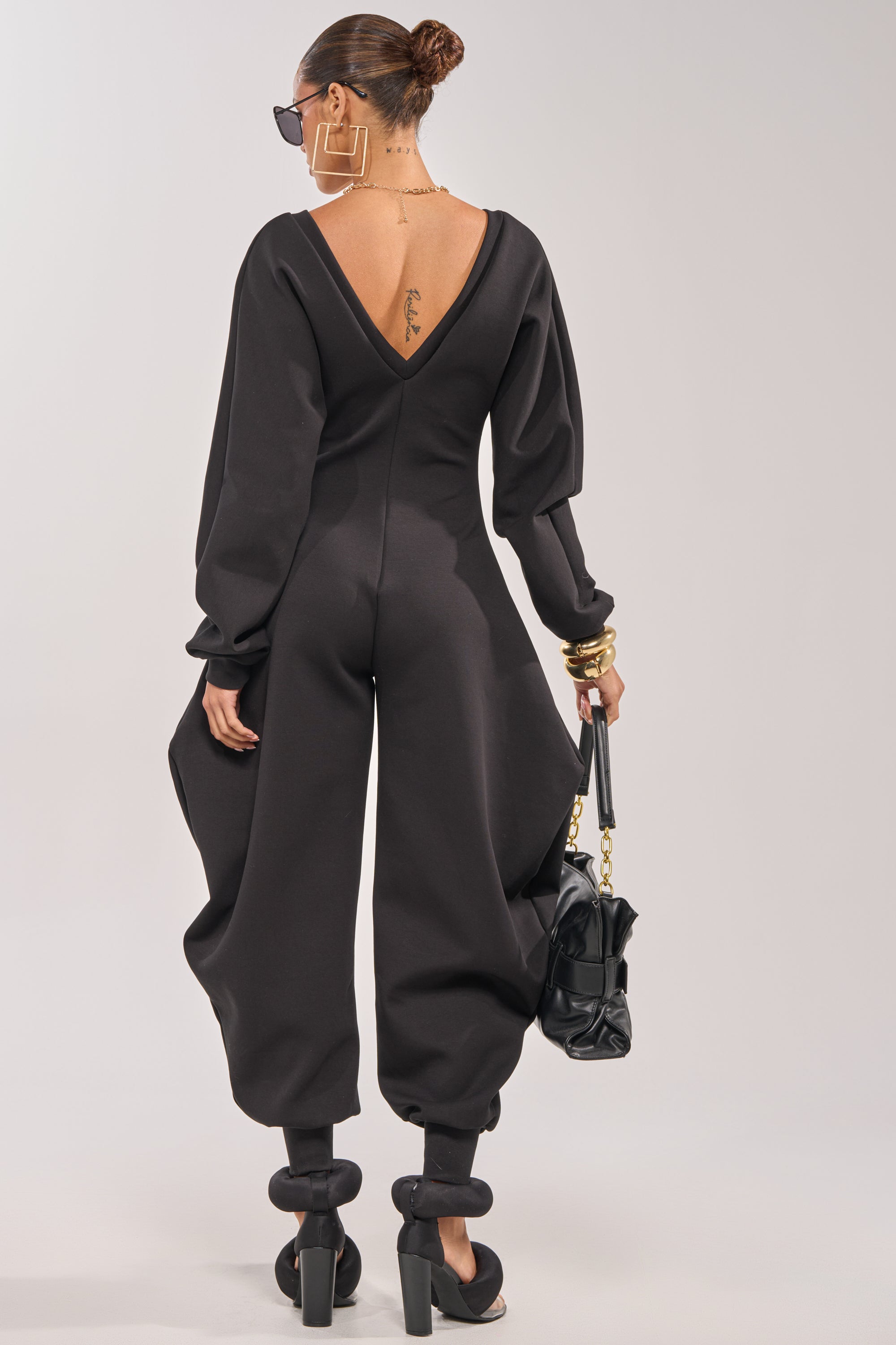 A person with a low bun and large hoop earrings wears the COMFY COZY SWEATSHIRT JUMPSUIT IN BLACK— a relaxed, long-sleeve V-neck style with billowed legs—paired with a black handbag, heels featuring spherical details, and showcasing a back tattoo. 2