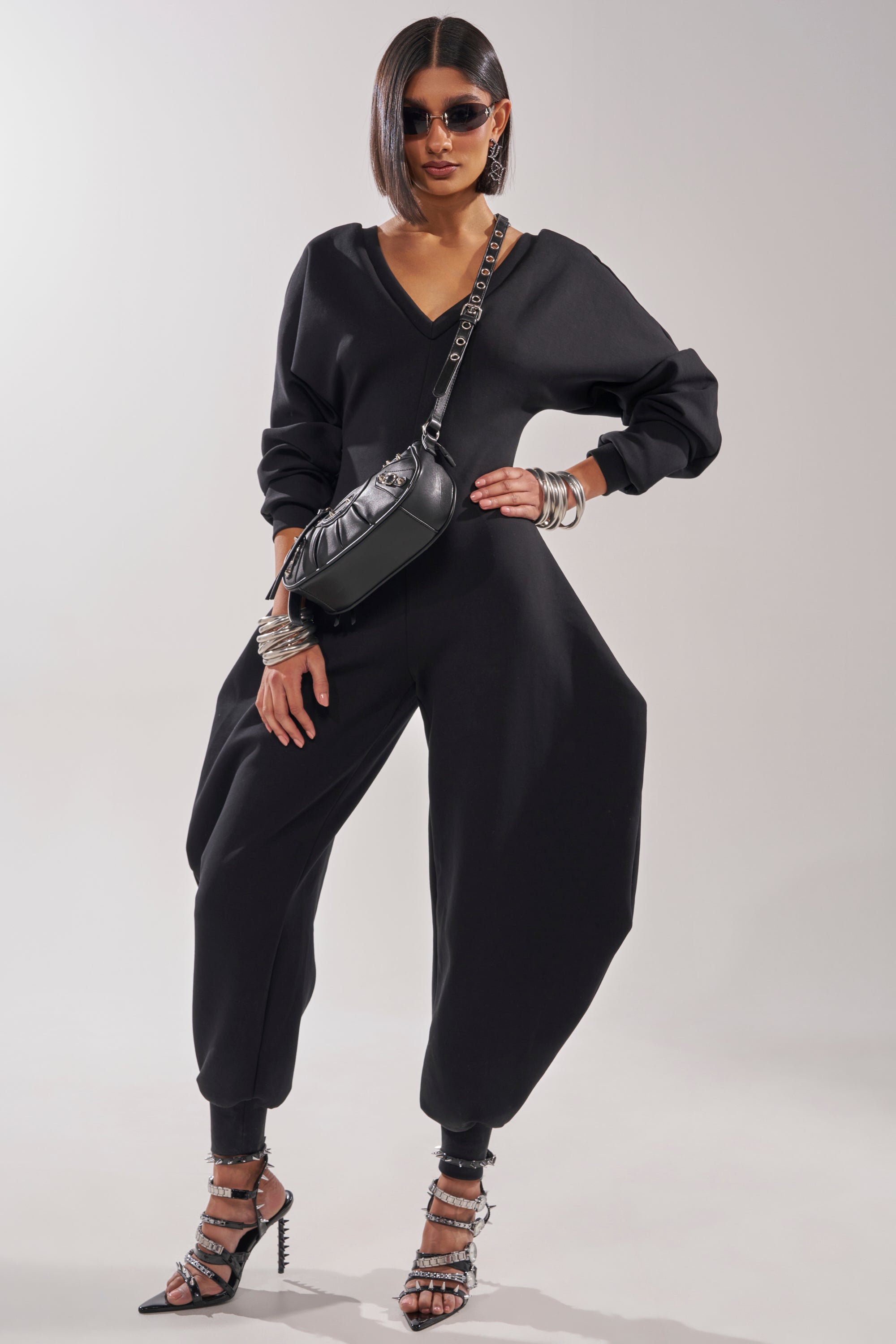 A woman exudes confidence in the COMFY COZY SWEATSHIRT JUMPSUIT IN BLACK, featuring a V-neck and stretchy cotton fabric. She styles it with strappy heels, silver bangles, sunglasses, and a black crossbody bag against a light backdrop. 2
