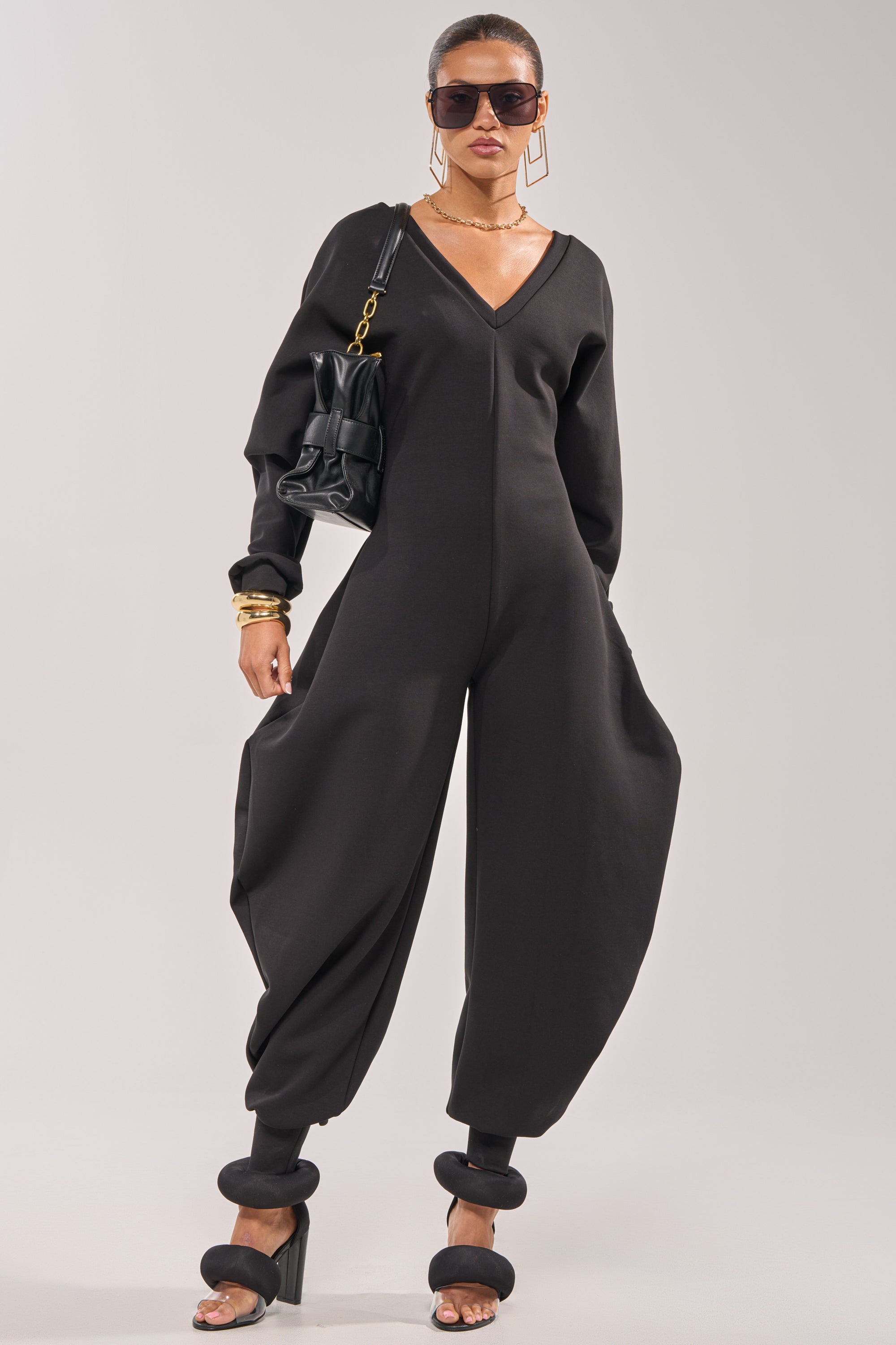 A woman stands confidently in the COMFY COZY SWEATSHIRT JUMPSUIT IN BLACK, styled with black heeled sandals, sunglasses, gold jewelry, and a black handbag with a gold chain strap against a plain light background. 3