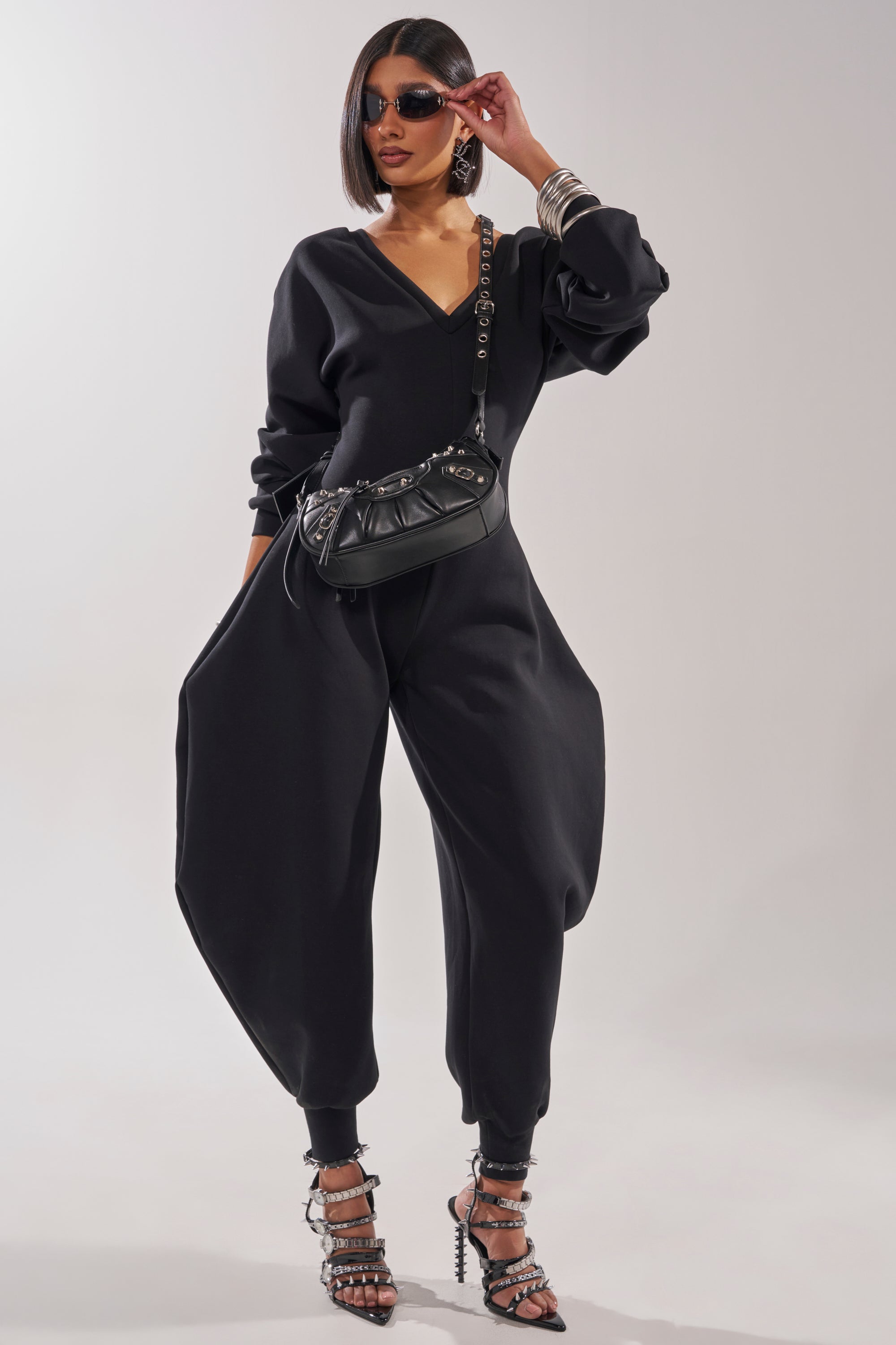A woman wears the COMFY COZY SWEATSHIRT JUMPSUIT IN BLACK with wide legs, strappy black heels, sunglasses, bracelets, and carries a black shoulder bag while posing confidently against a plain background. 3