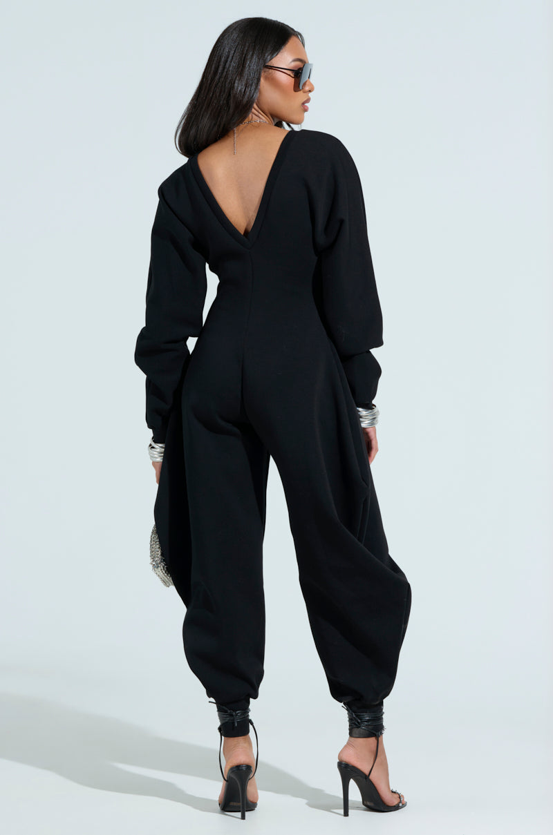 A woman in a stylish black COMFY COZY SWEATSHIRT JUMPSUIT stands facing away, accessorized with sunglasses, high heels, and bracelets on both wrists. She holds a textured silver clutch against a pale blue background that highlights her chic style.