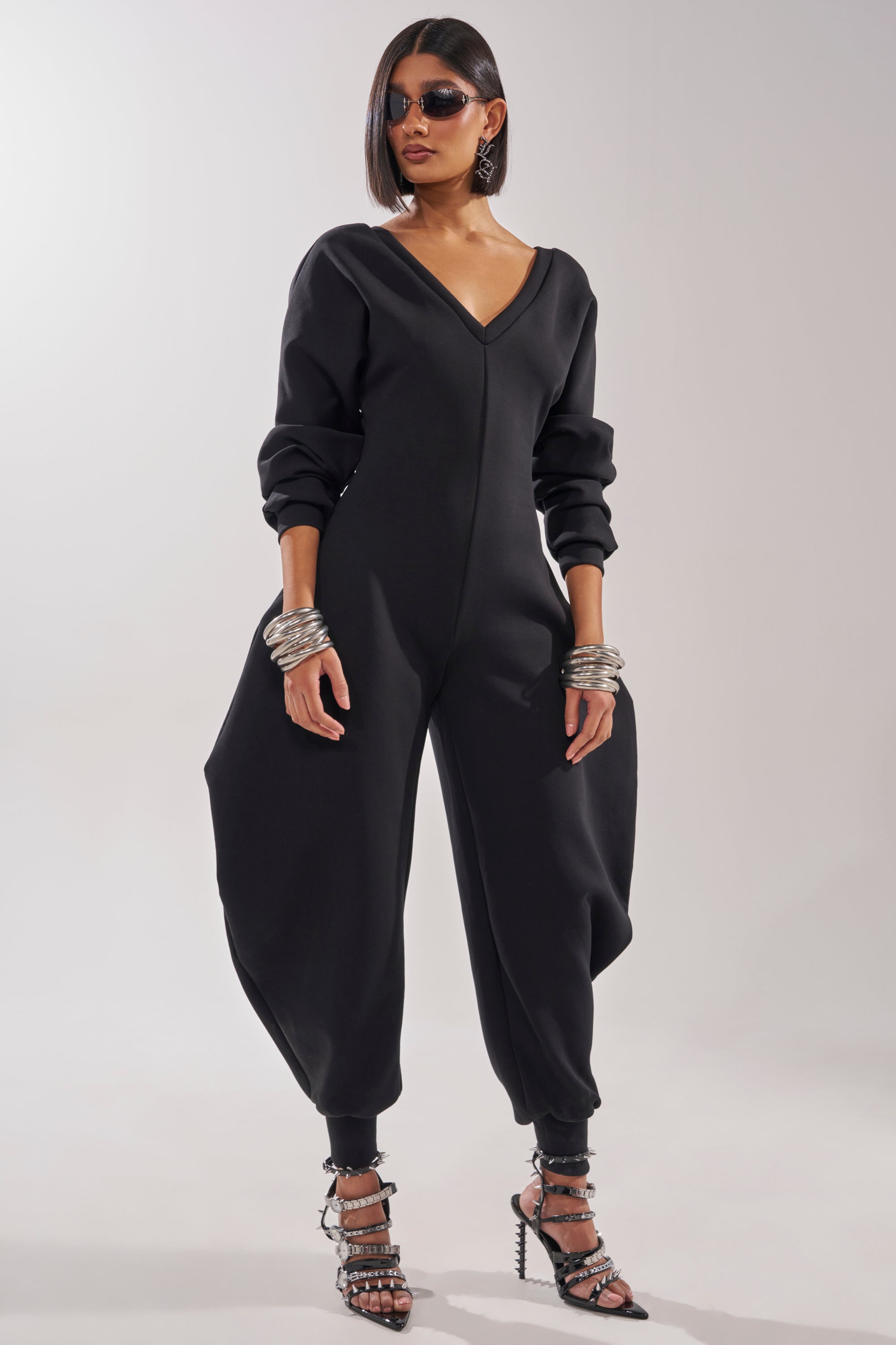 A woman confidently poses against a plain background in the COMFY COZY SWEATSHIRT JUMPSUIT IN BLACK, styled with silver bangles, strappy black heels, and sunglasses. 4