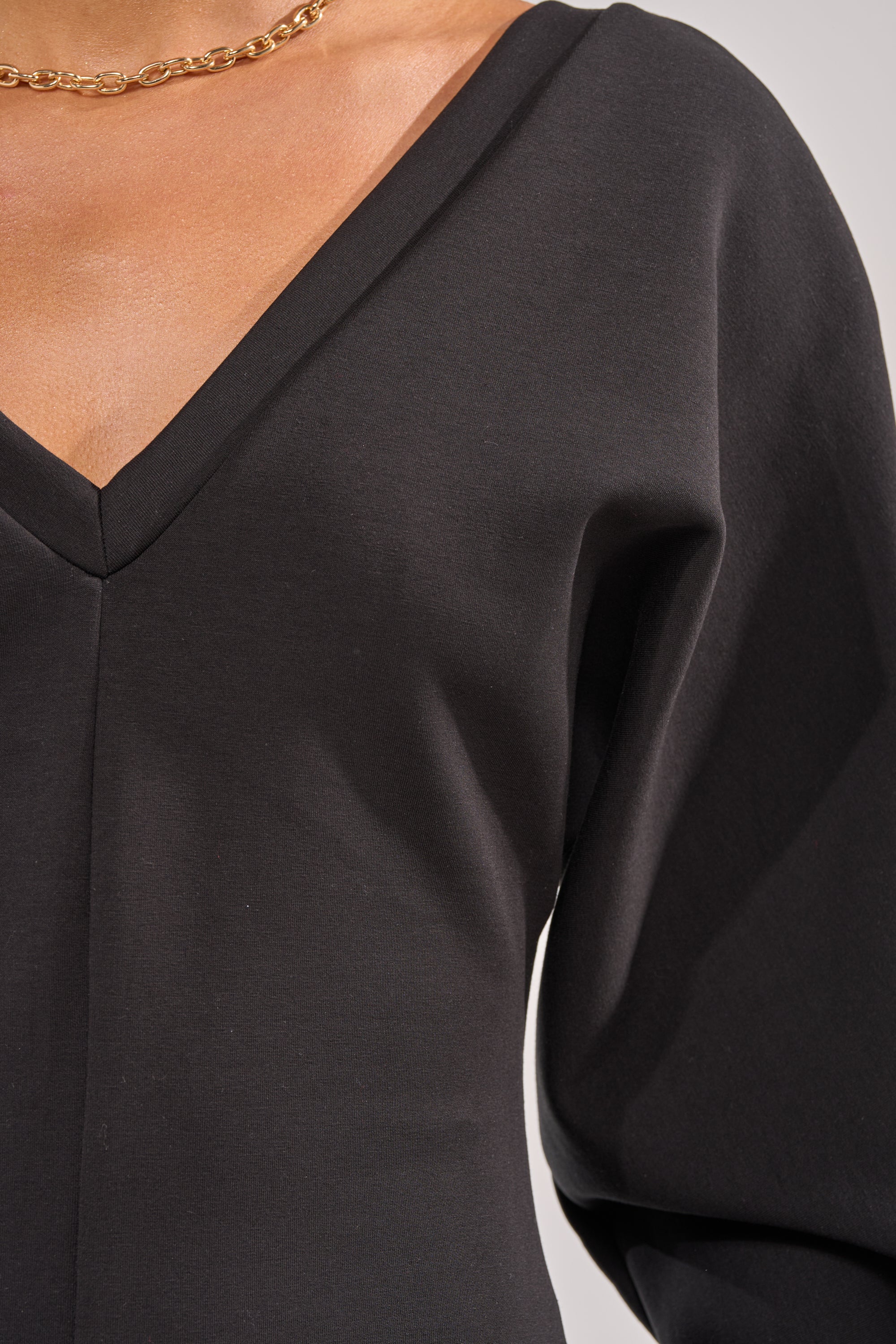 Close-up of a person wearing the COMFY COZY SWEATSHIRT JUMPSUIT IN BLACK, featuring a V-neck, long sleeves, and a gold chain necklace, with part of the chest and shoulder visible. 4