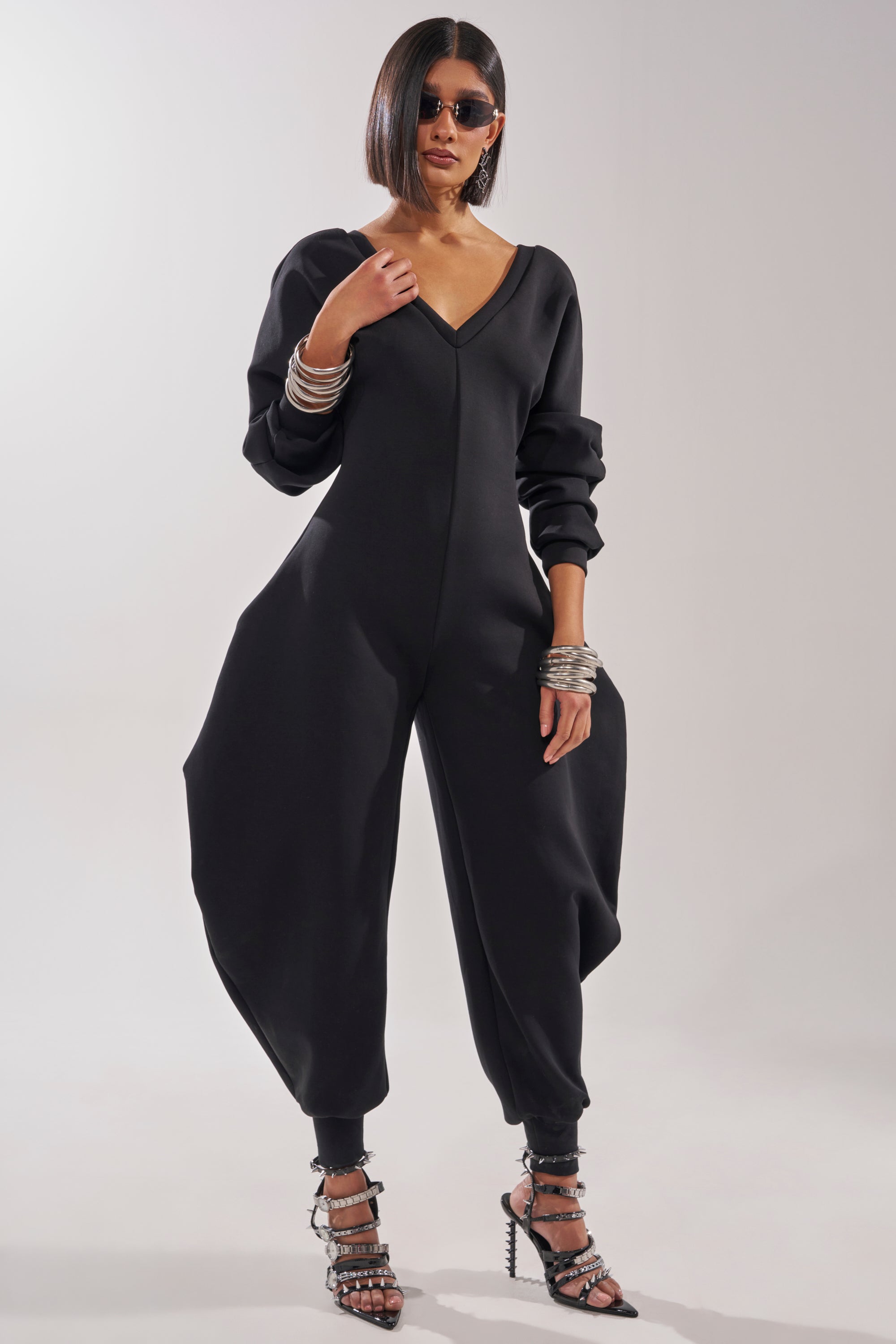 A woman in sunglasses sports the COMFY COZY SWEATSHIRT JUMPSUIT IN BLACK, featuring voluminous sculpted legs and long sleeves, styled with stacked silver bangles and strappy high-heeled sandals for extra flair. 5