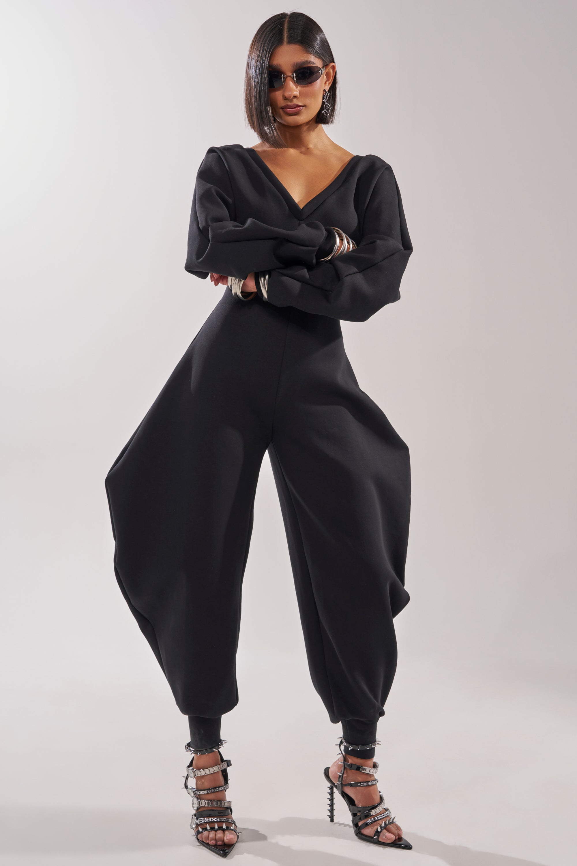 A woman with straight shoulder-length hair and sunglasses wears the COMFY COZY SWEATSHIRT JUMPSUIT IN BLACK, featuring voluminous legs. She stands confidently in strappy heels, arms folded, against a plain light background. 6