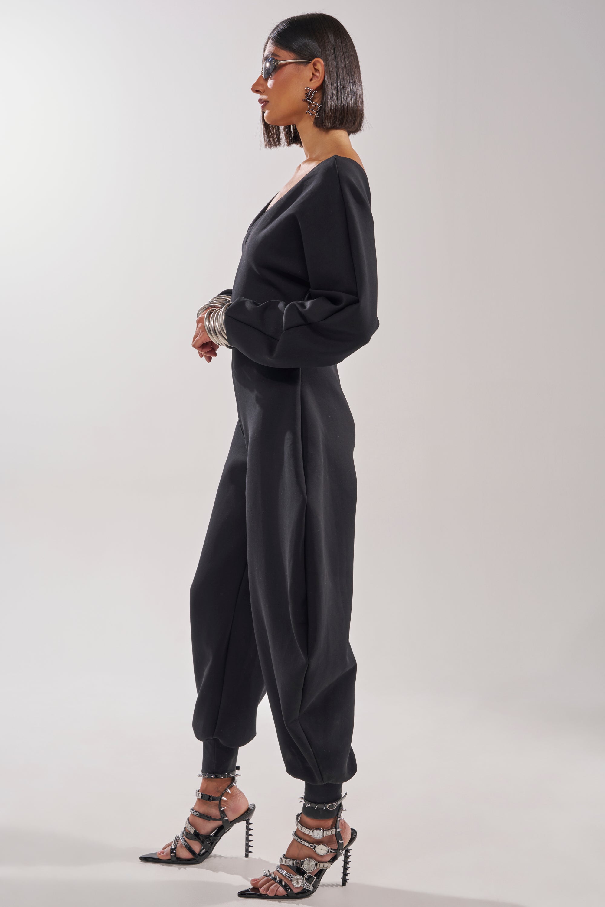 A woman with a sleek bob stands in profile wearing the COMFY COZY SWEATSHIRT JUMPSUIT IN BLACK, paired with statement earrings, silver bracelets, sunglasses, and black strappy high heels against a plain light background. 7
