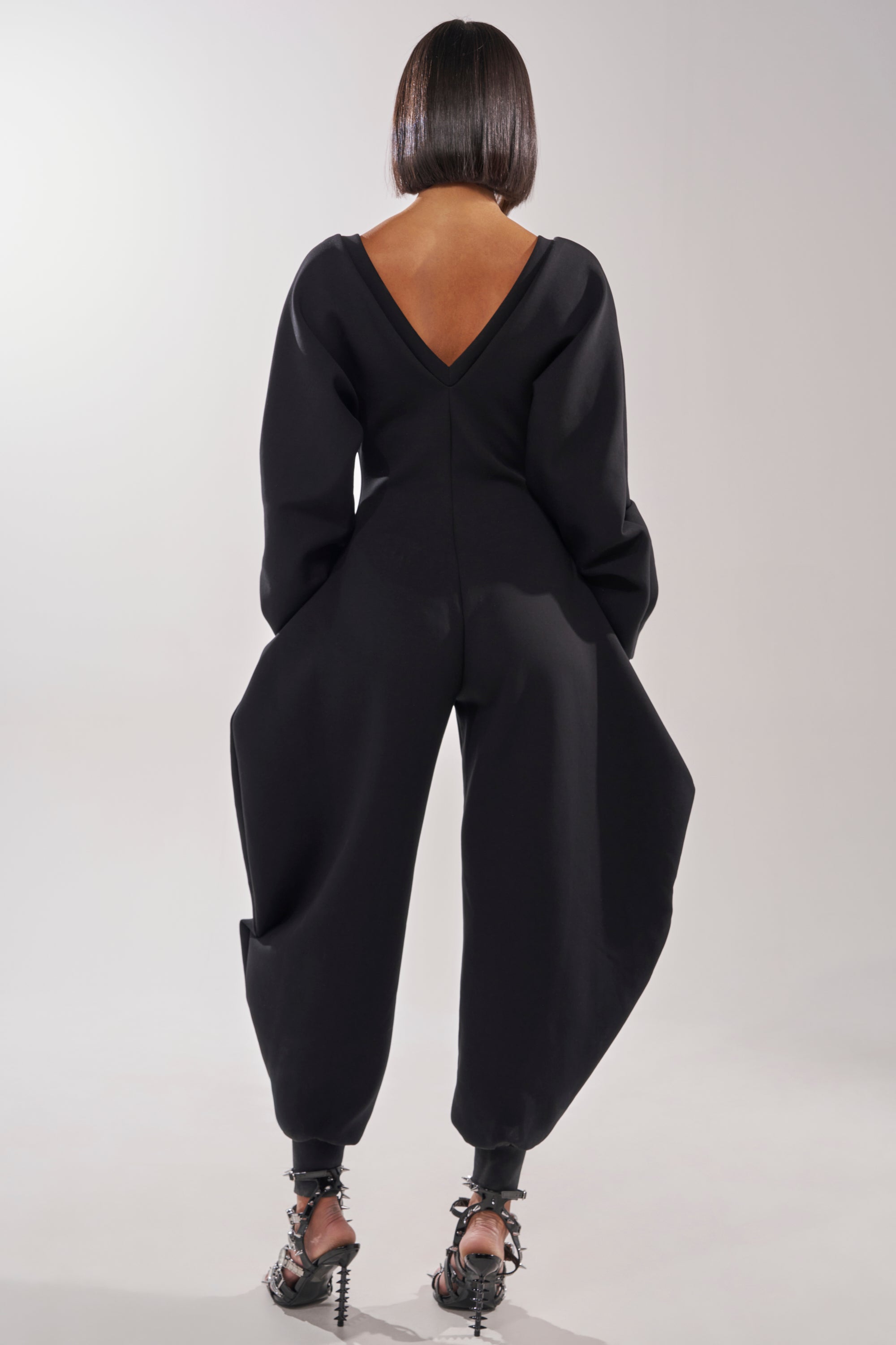A person with a sleek bob stands facing away, wearing the COMFY COZY SWEATSHIRT JUMPSUIT IN BLACK featuring a deep V-back, voluminous legs, and long sleeves. They complete the look with black open-toe heels and ankle straps. 8