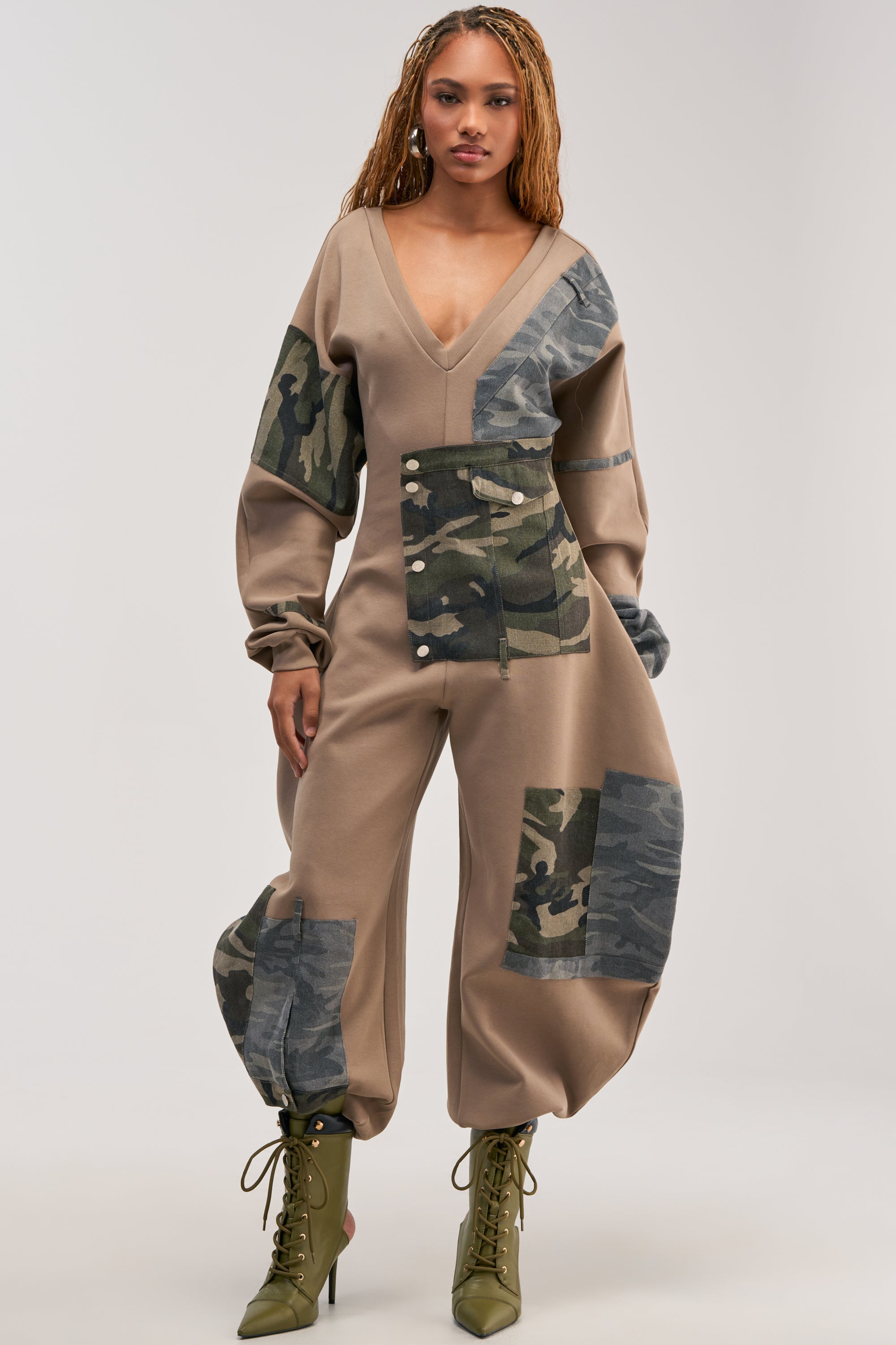 A woman models the COMFY COZY PATCHWORK JUMPSUIT IN CAMO—featuring tan and camo details, loose legs, and long puffed sleeves—for an effortlessly cool streetwear loungewear look. She pairs it with green lace-up boots against a neutral background.