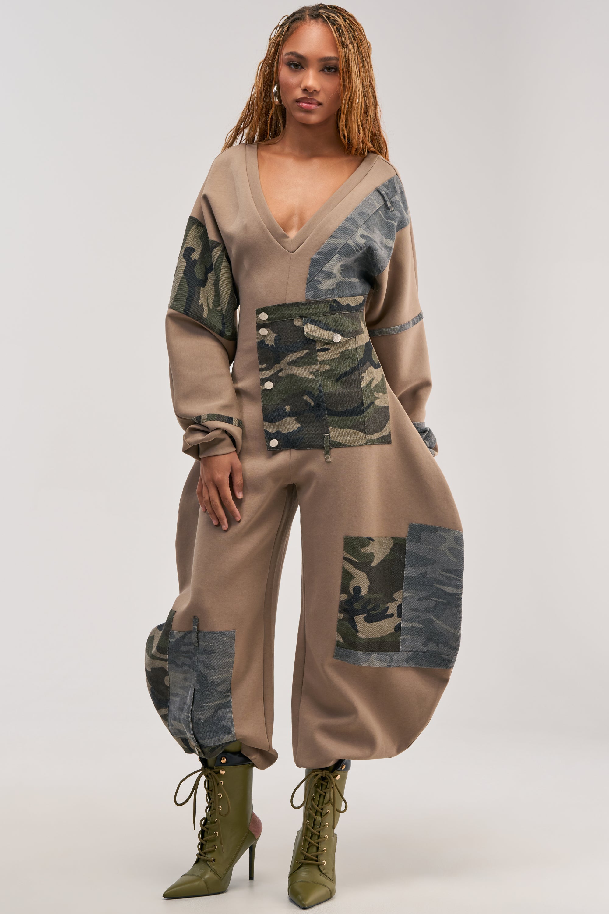 Wearing the COMFY COZY PATCHWORK JUMPSUIT IN CAMO—with its olive camo patchwork details, deep V-neckline, and loose, tapered legs—a woman pairs it with green lace-up heeled boots as she stands against a plain light background, effortlessly embodying streetwear-meets-loungewear style.