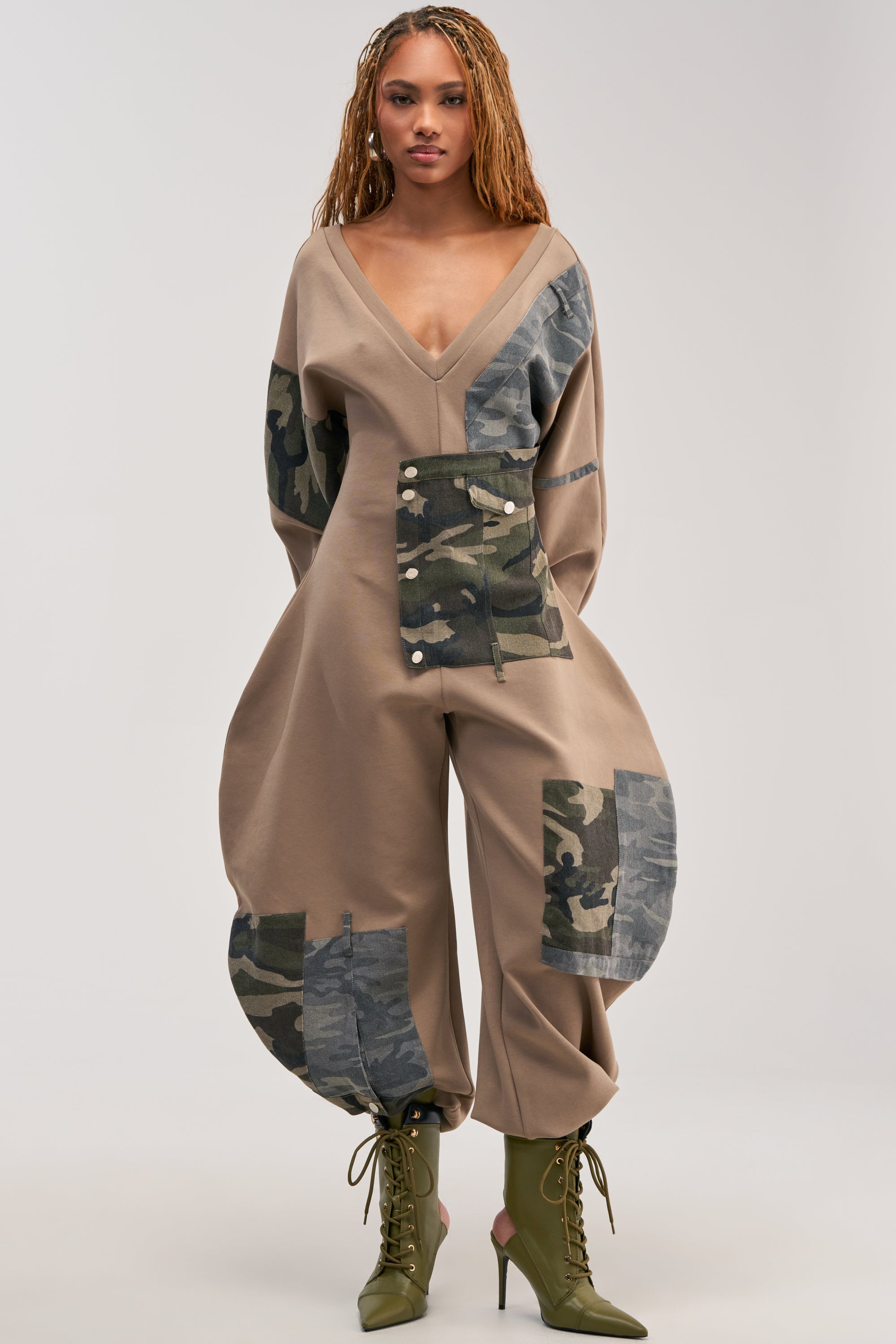 A person wearing the COMFY COZY PATCHWORK JUMPSUIT IN CAMO—an urban streetwear-inspired jumpsuit with tan fabric, olive camo patchwork, a deep V-neckline, wide long sleeves, and ballooned pant legs. They pair it with green lace-up boots and stand against a plain light background.