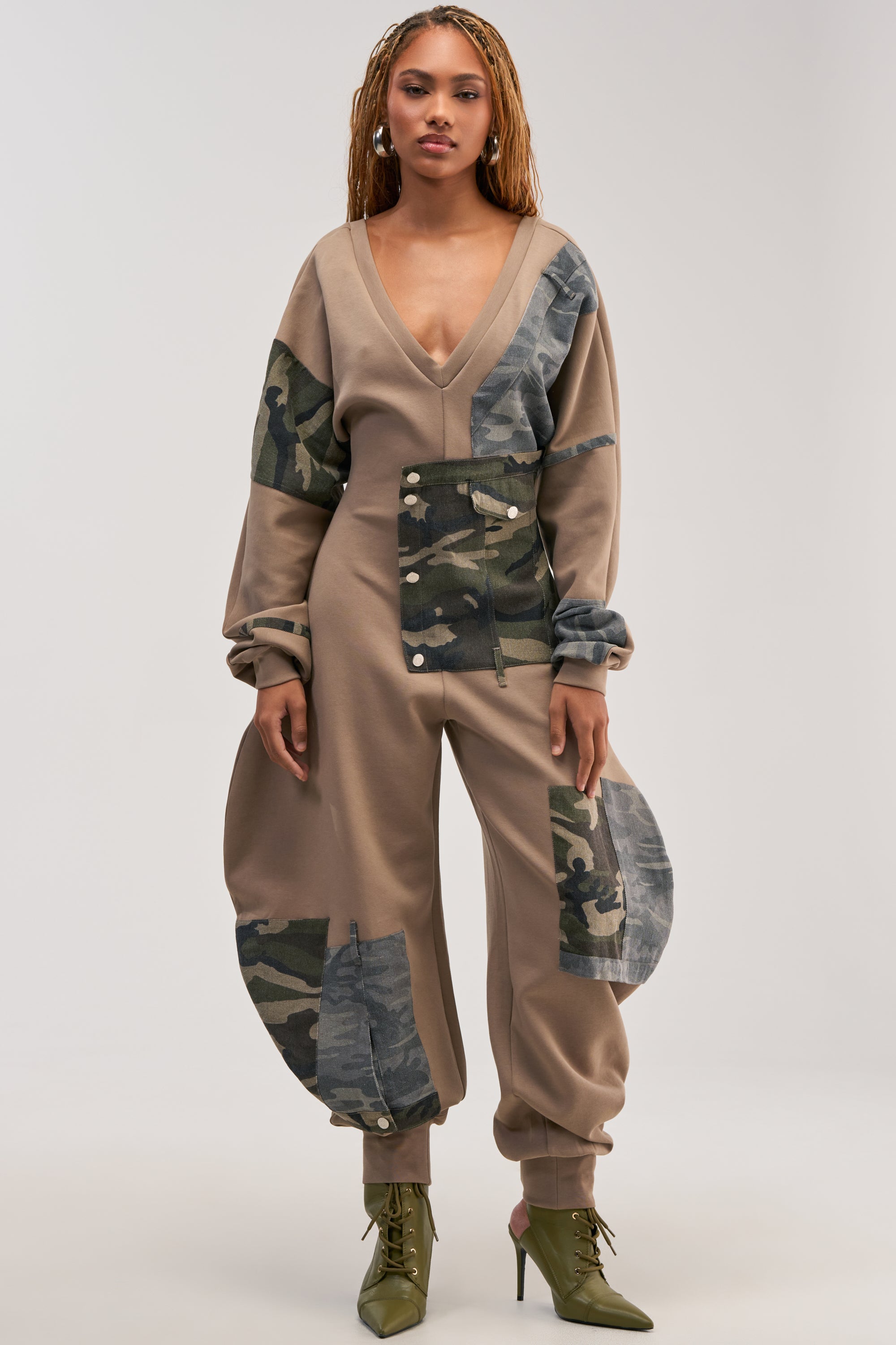 A woman stands facing forward against a neutral background, modeling the COMFY COZY PATCHWORK JUMPSUIT IN CAMO. The jumpsuit features green camouflage patchwork, a deep V-neckline, wide rounded side panels, and long sleeves for a modern streetwear-inspired loungewear look. She completes the outfit with green lace-up boots and wears her hair in braids.