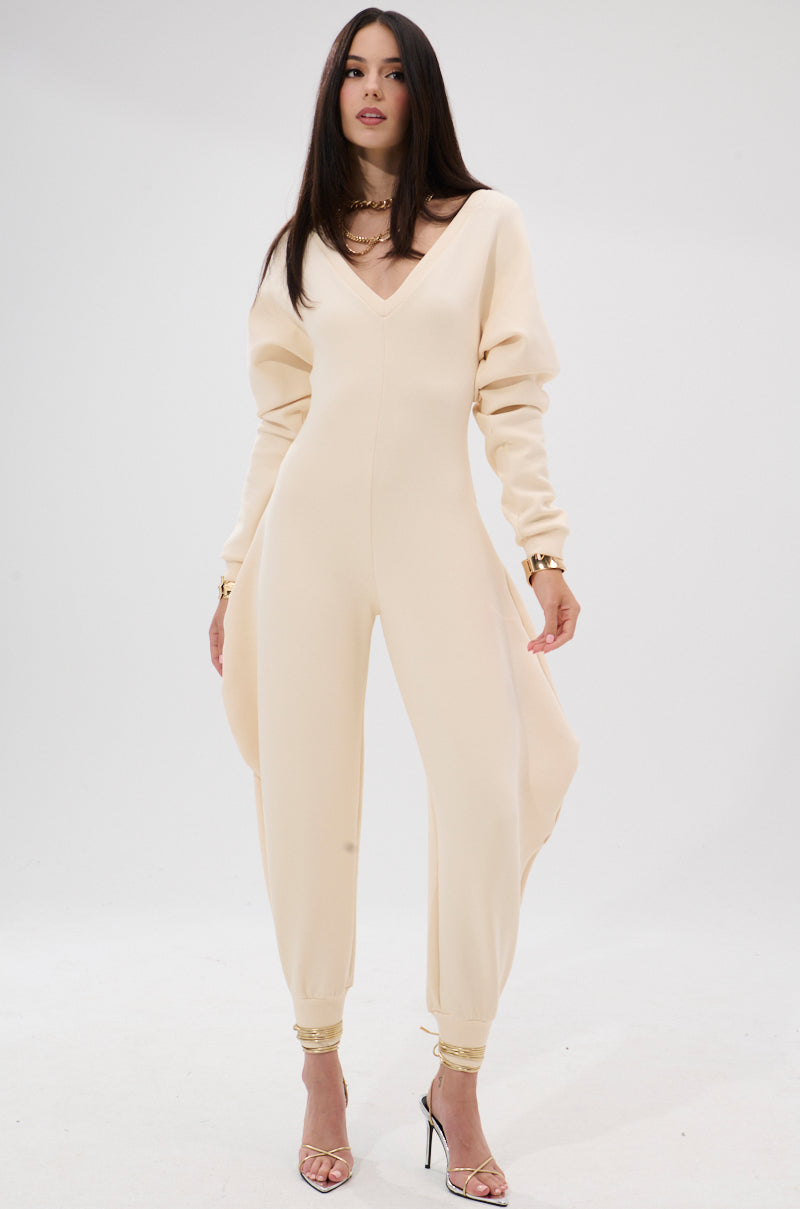 A woman stands against a white background wearing the COMFY COZY SWEATSHIRT JUMPSUIT IN BONE—stretchy, long-sleeved, with draped wide legs—styled with gold jewelry and clear heels for an effortlessly elegant loungewear look.