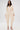 A woman stands against a white background wearing the COMFY COZY SWEATSHIRT JUMPSUIT IN BONE—stretchy, long-sleeved, with draped wide legs—styled with gold jewelry and clear heels for an effortlessly elegant loungewear look.