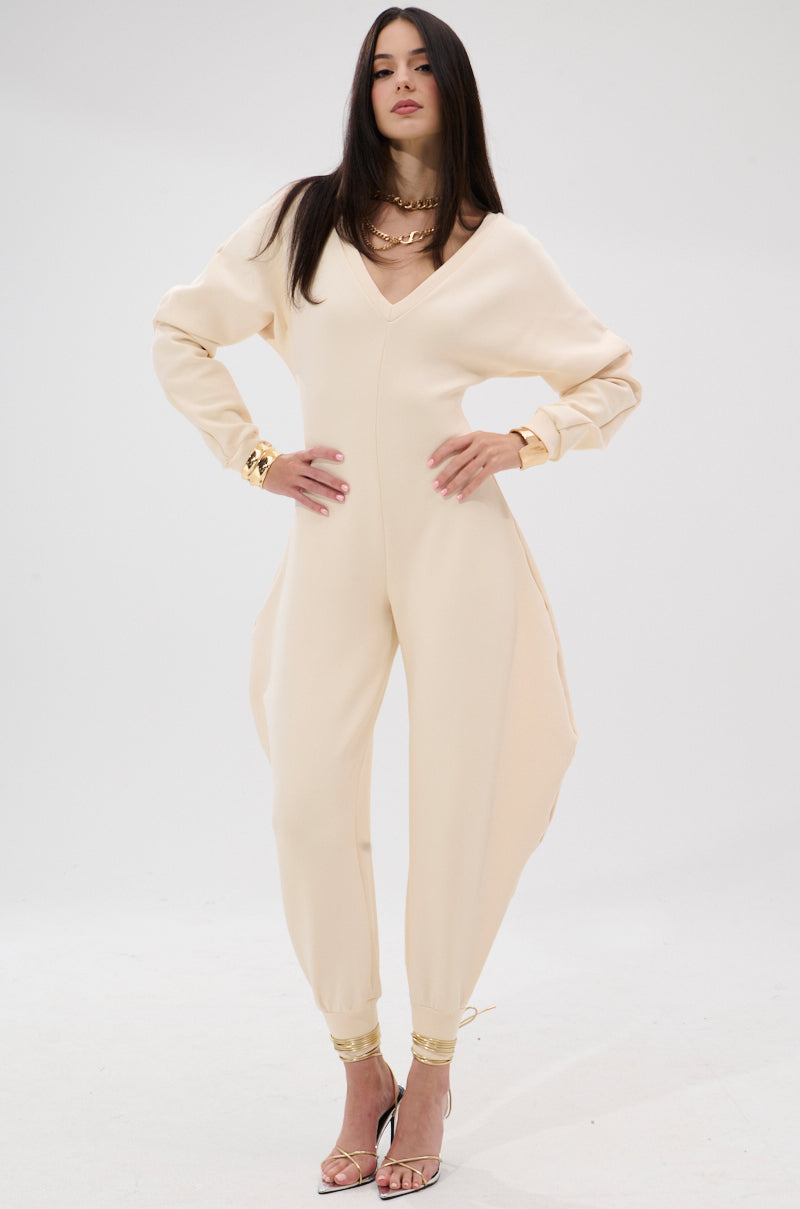 A woman stands confidently in the COMFY COZY SWEATSHIRT JUMPSUIT IN BONE—featuring long sleeves, a V-neck, and gathered ankles—styled with gold jewelry and heeled sandals against a white background.