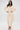 A woman stands confidently in the COMFY COZY SWEATSHIRT JUMPSUIT IN BONE—featuring long sleeves, a V-neck, and gathered ankles—styled with gold jewelry and heeled sandals against a white background.