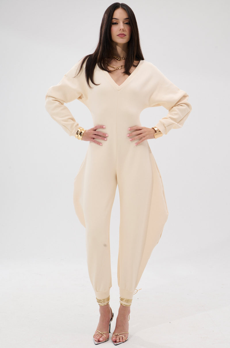 A woman stands with hands on hips against a white background, wearing the COMFY COZY SWEATSHIRT JUMPSUIT IN BONE—a cream-colored, stretchy cotton jumpsuit with a deep V-neckline—paired with gold heels.