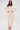 A woman stands with hands on hips against a white background, wearing the COMFY COZY SWEATSHIRT JUMPSUIT IN BONE—a cream-colored, stretchy cotton jumpsuit with a deep V-neckline—paired with gold heels.