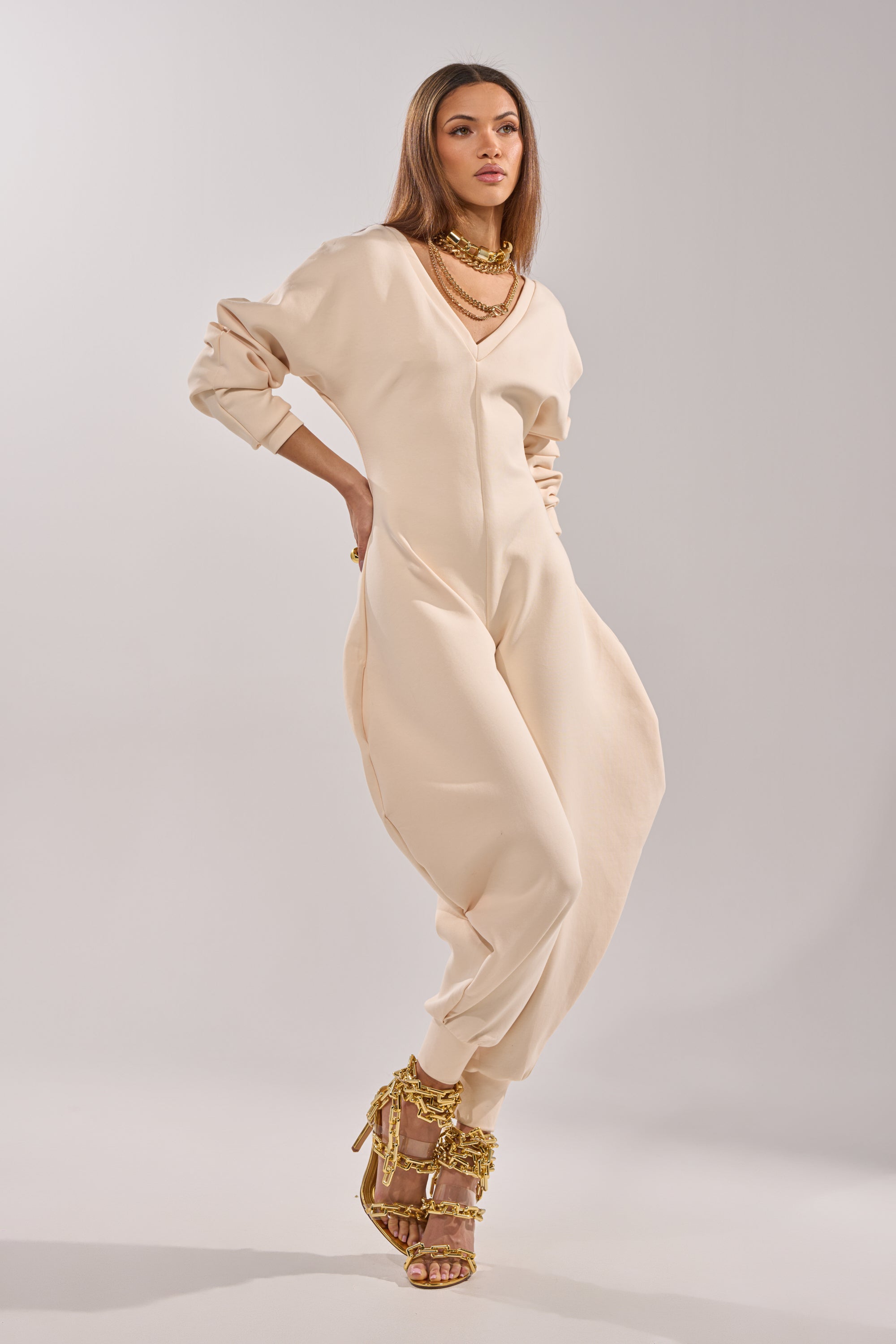 A woman poses confidently in the COMFY COZY SWEATSHIRT JUMPSUIT IN BONE, featuring stretchy cotton, long sleeves, and cuffed ankles, styled with gold chain necklaces and matching heels against a plain light background. 2