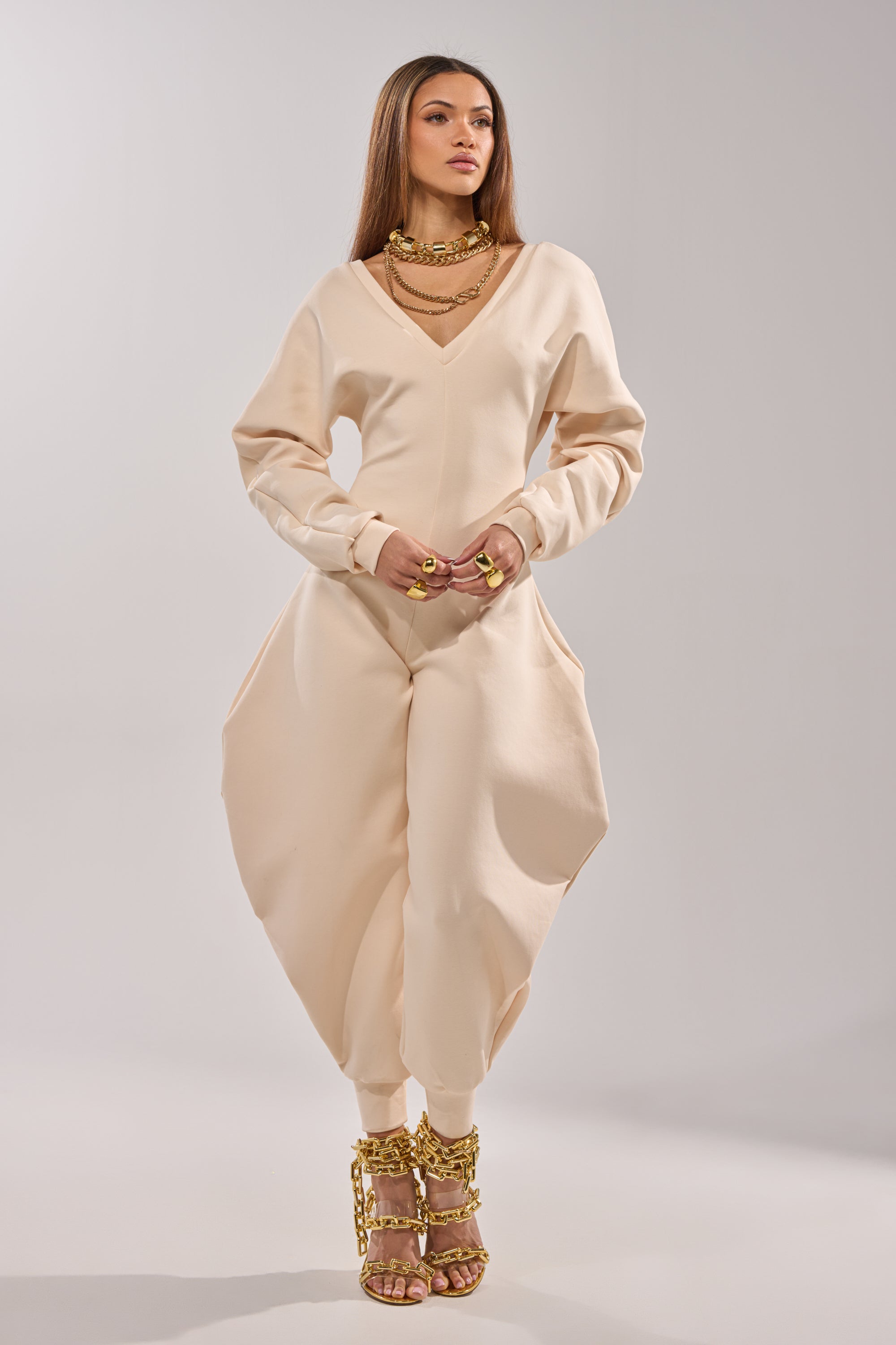 A woman models the COMFY COZY SWEATSHIRT JUMPSUIT IN BONE, featuring voluminous legs and soft stretch cotton. She accessorizes with layered gold jewelry and sandals with chunky gold chains against a plain light background. 3