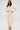 A woman poses confidently in the COMFY COZY SWEATSHIRT JUMPSUIT IN BONE, featuring loose, draped sides, long sleeves, and a V-neck. She accessorizes with gold jewelry and heels against a white background for a chic loungewear look.
