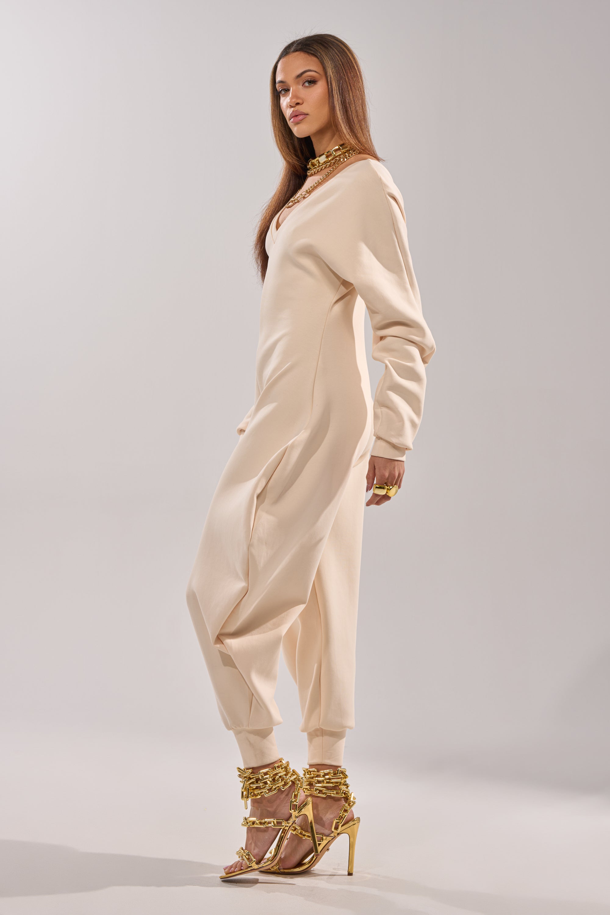 A woman stands in profile against a plain light background, wearing the COMFY COZY SWEATSHIRT JUMPSUIT IN BONE, styled with gold statement jewelry and gold strappy high-heeled sandals. 4