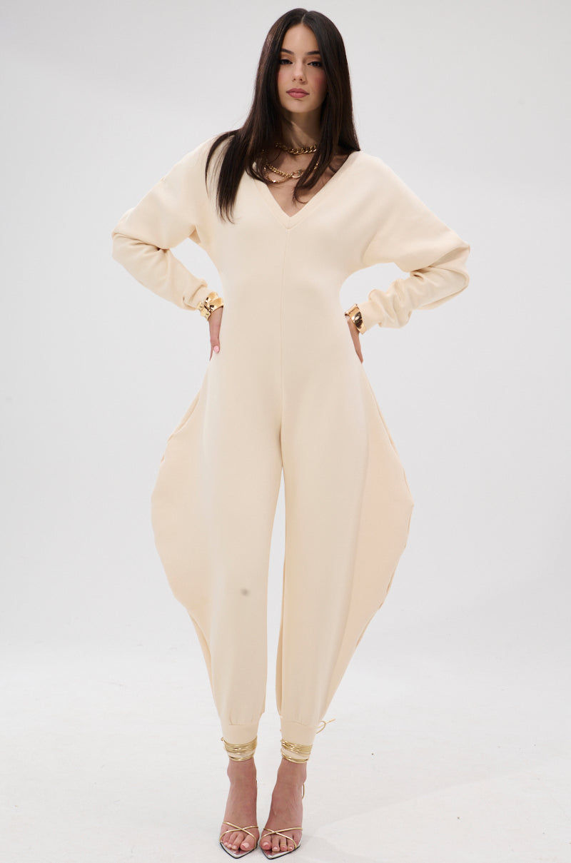 A woman models the COMFY COZY SWEATSHIRT JUMPSUIT IN BONE against a white backdrop. The stretchy, sculpted design with fitted ankles is perfect for lounging, styled here with straight hair, gold jewelry, and open-toe heels.