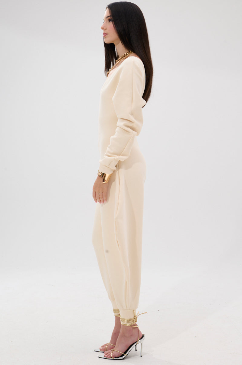 A woman stands in profile against a white background, wearing the COMFY COZY SWEATSHIRT JUMPSUIT IN BONE, styled with gold ankle cuffs, gold jewelry, and strappy high-heeled sandals.
