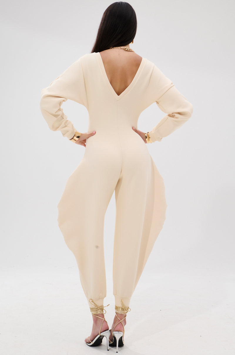 A woman faces away in the COMFY COZY SWEATSHIRT JUMPSUIT IN BONE, featuring a deep V-back, loose sleeves, and wide legs. Styled with strappy heels and gold jewelry, she stands against a light background for an elevated loungewear look.