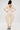 A woman faces away in the COMFY COZY SWEATSHIRT JUMPSUIT IN BONE, featuring a deep V-back, loose sleeves, and wide legs. Styled with strappy heels and gold jewelry, she stands against a light background for an elevated loungewear look.