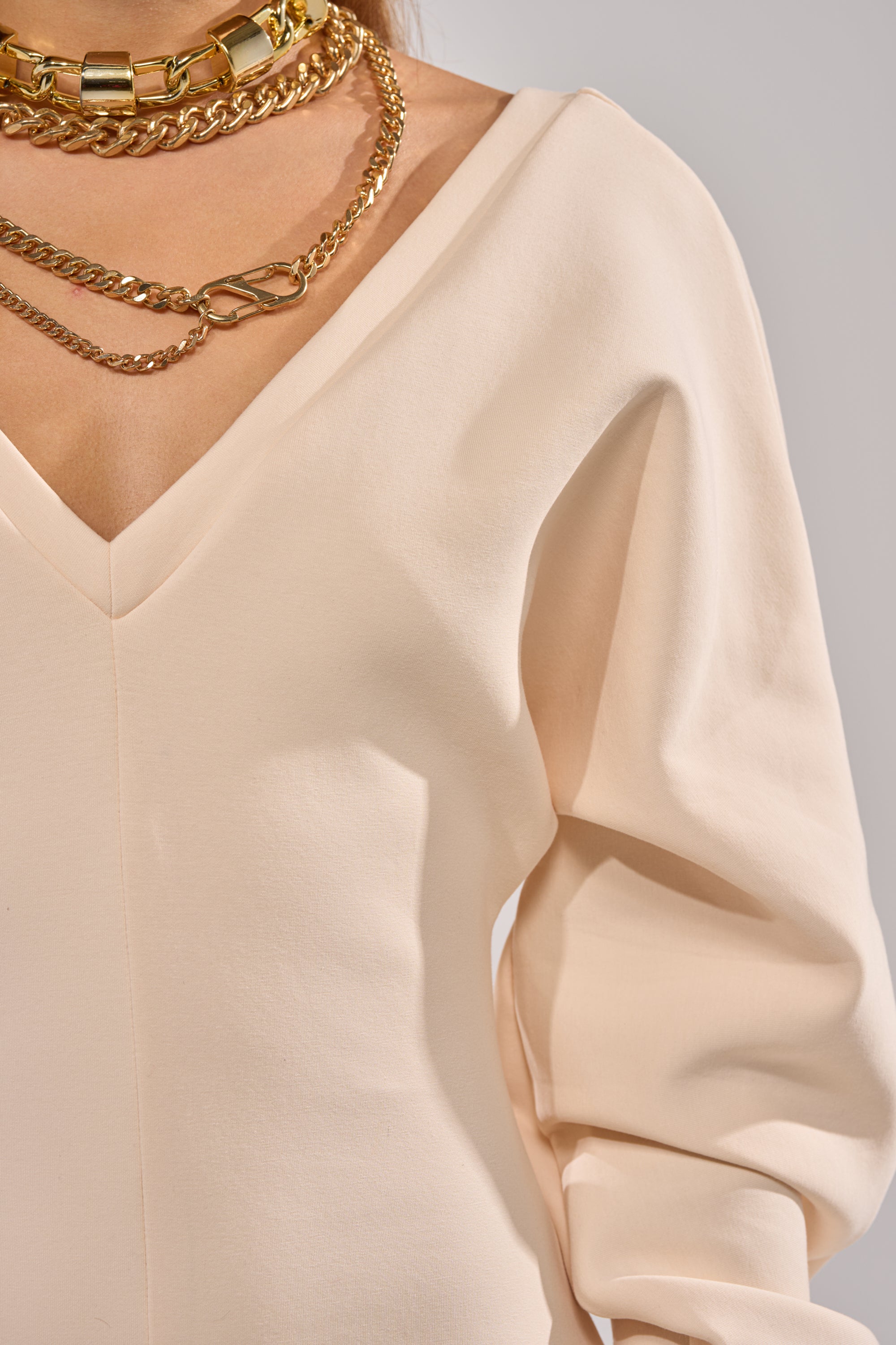 A person wearing the COMFY COZY SWEATSHIRT JUMPSUIT IN BONE, styled with layered gold chain necklaces. The cropped photo highlights the upper torso and neck, showcasing both the soft loungewear and chic accessories. 6