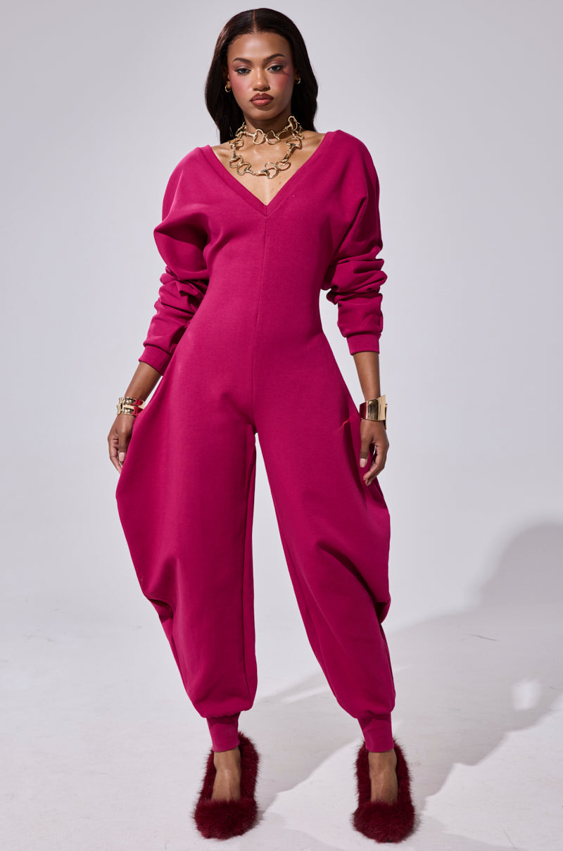 A woman exudes confidence in the COMFY COZY SWEATSHIRT JUMPSUIT IN PLUM, featuring voluminous, tapered legs and long sleeves. Paired with magenta heels and gold jewelry, she embraces a comfy-cozy style against a simple light gray background.