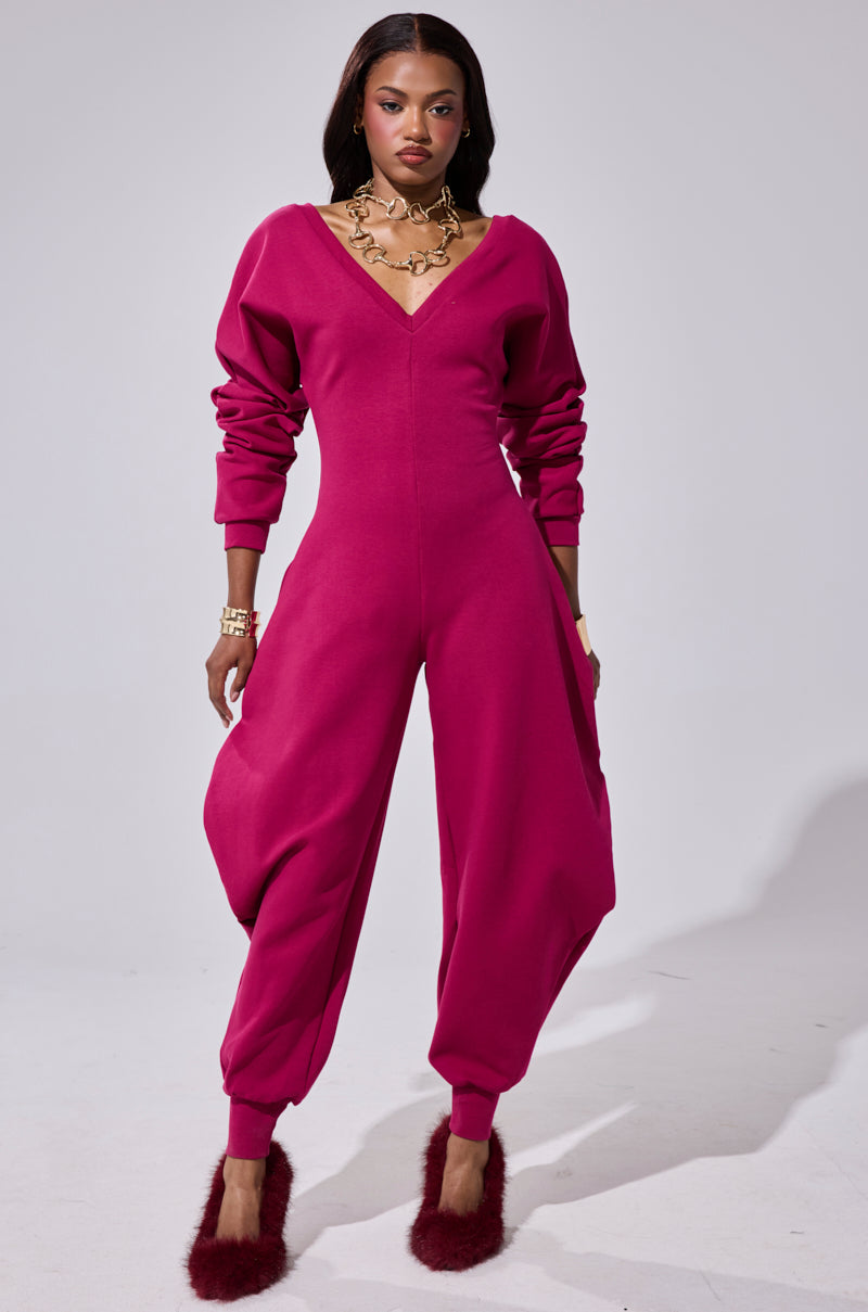 A woman stands confidently in the COMFY COZY SWEATSHIRT JUMPSUIT IN PLUM, featuring bold, voluminous legs and fitted cuffs. She accessorizes with gold jewelry and fuzzy burgundy heels, set against a plain light background.
