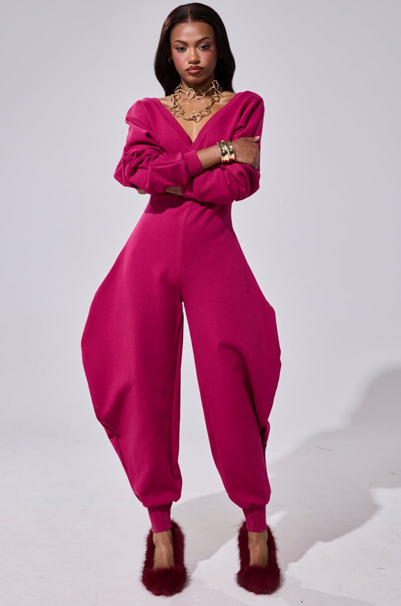 A woman stands confidently in the COMFY COZY SWEATSHIRT JUMPSUIT IN PLUM with voluminous legs, long sleeves, and a V-neckline, accessorized with a chunky gold necklace, bracelet, and fuzzy pink slippers.