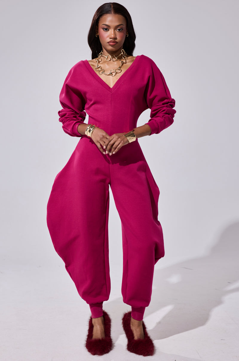 A woman stands confidently in the COMFY COZY SWEATSHIRT JUMPSUIT IN PLUM, featuring sculpted hips and long sleeves. She pairs it with gold jewelry and red fuzzy slippers, posing against a plain white background.