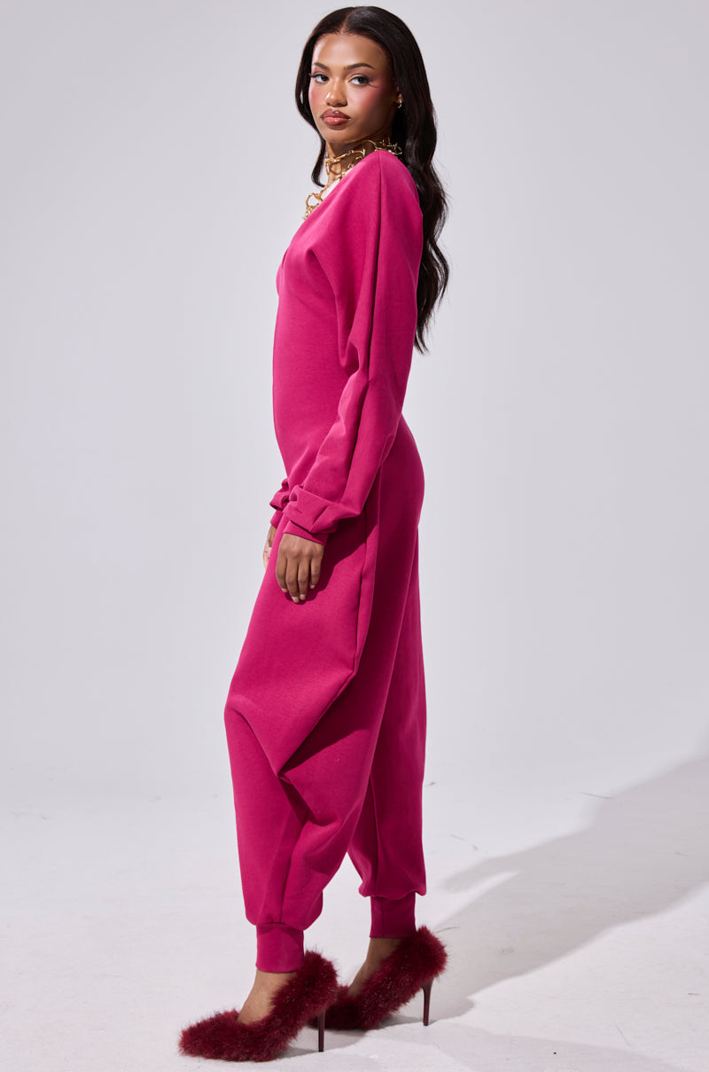 A woman with long dark hair and gold jewelry stands sideways in the COMFY COZY SWEATSHIRT JUMPSUIT IN PLUM, featuring jogger cuffs. She pairs it with fuzzy red heels and poses against a plain light background, wearing a neutral expression.