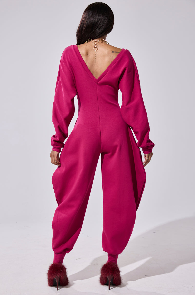 A person with long dark hair faces away, wearing the COMFY COZY SWEATSHIRT JUMPSUIT IN PLUM featuring a deep V-back, balloon sleeves, and billowy pant legs, paired with fluffy red high-heeled shoes.